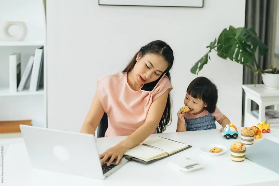 Working mother using laptop and taking care of child in home office with snacks and toys.