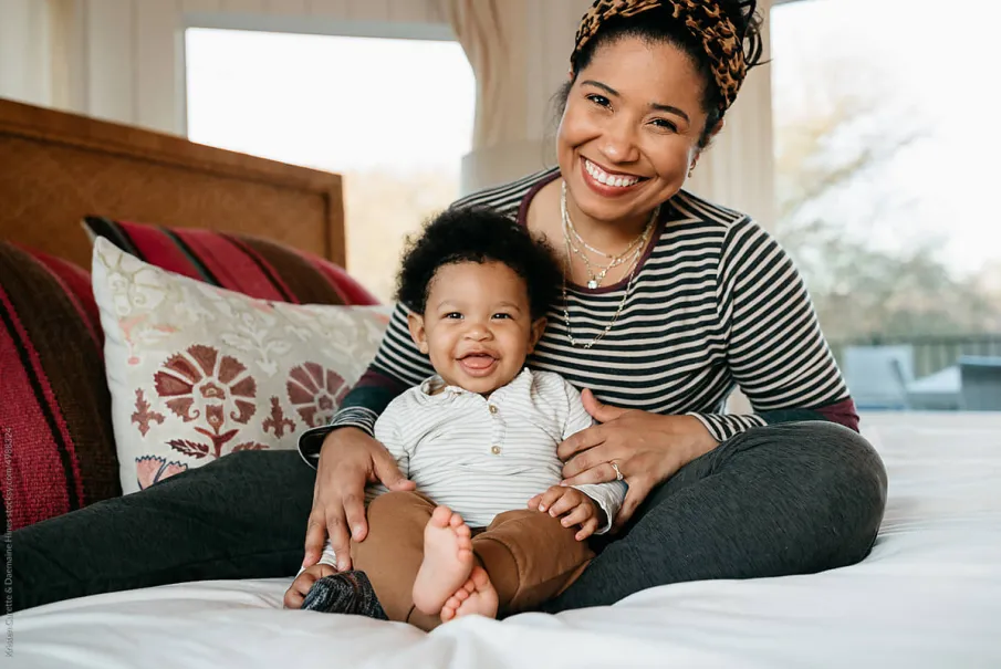 Smiling mother and baby sitting on a bed with decorative pillows.